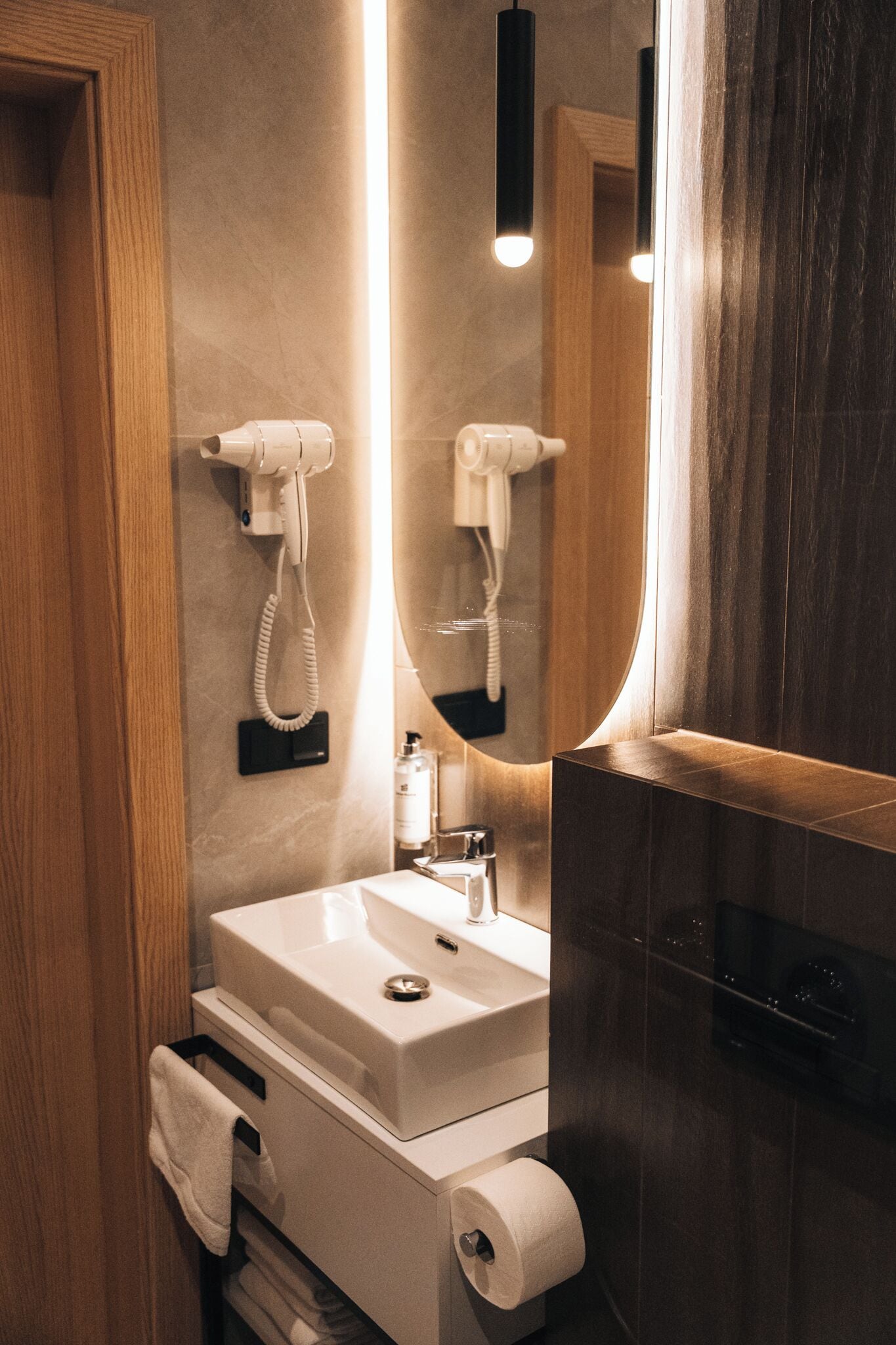 Un baño moderno con puerta de madera, lavamanos blanco, espejo y secador de pelo montado en la pared.