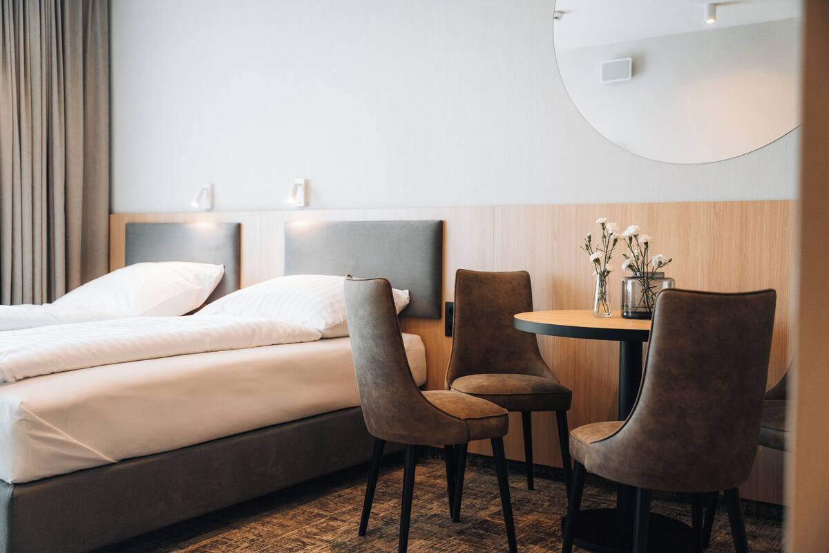 A hotel room with a bed, two chairs, a small table with a vase of flowers, and a round mirror on the wall.