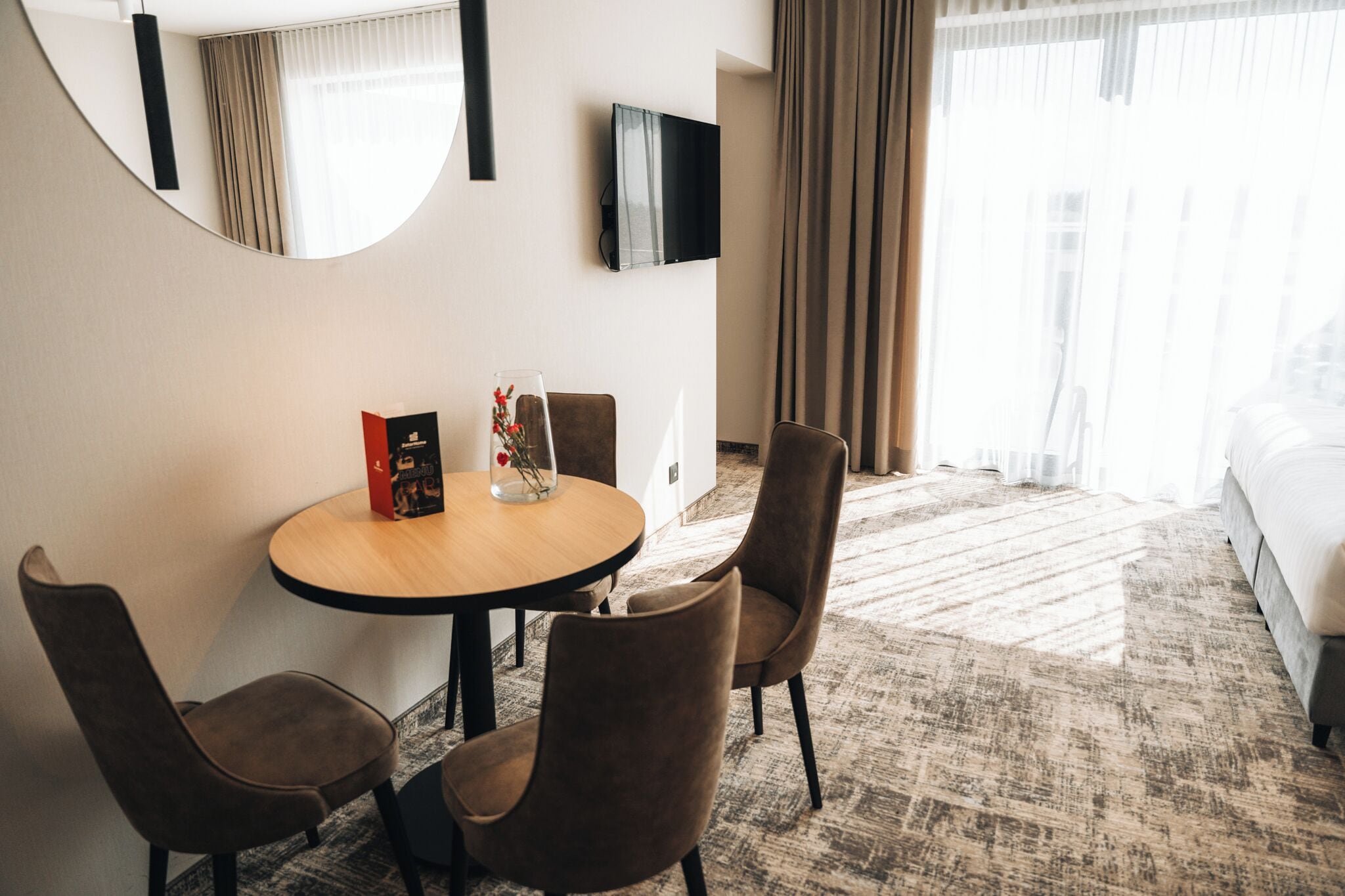 A hotel room with a round table, two chairs, a TV mounted on the wall, and a bed.