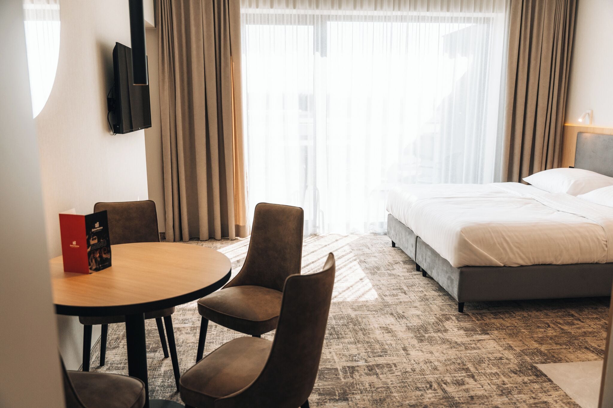A hotel room with a bed, a round table with chairs, a TV mounted on the wall, and a window with curtains.