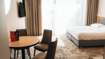 A hotel room with a bed, a round table with chairs, a TV mounted on the wall, and a window with curtains.