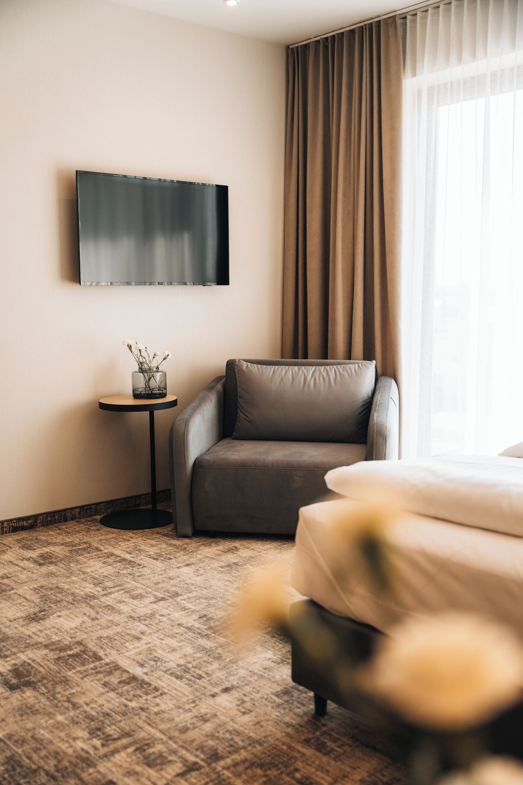 Een moderne hotelkamer met een bank, een klein bijzettafeltje met een vaas, een flatscreen televisie aan de muur en een groot raam met gordijnen.