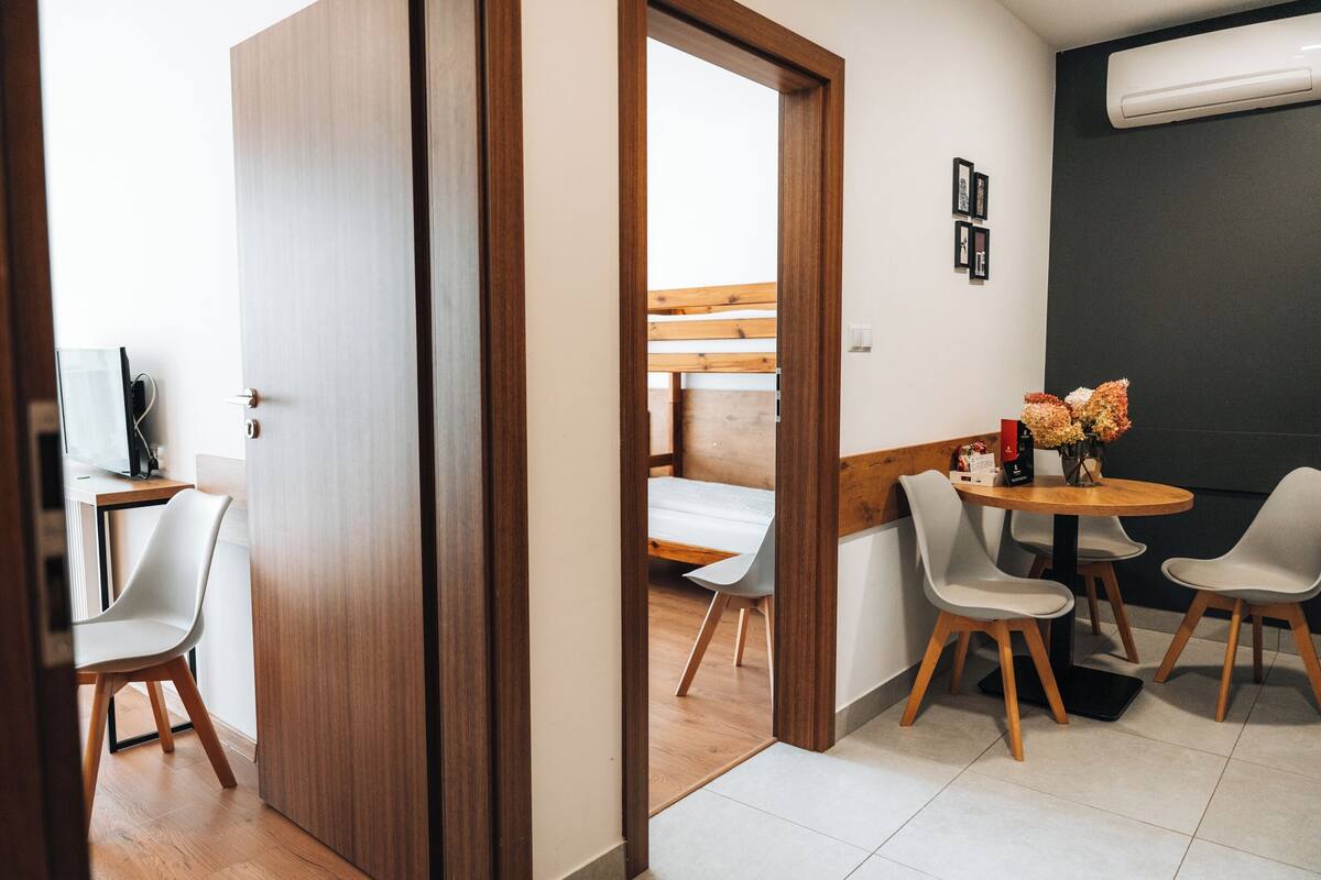 A modern room with a wooden door, a small dining area with a round table and two chairs, and a bed in the background.