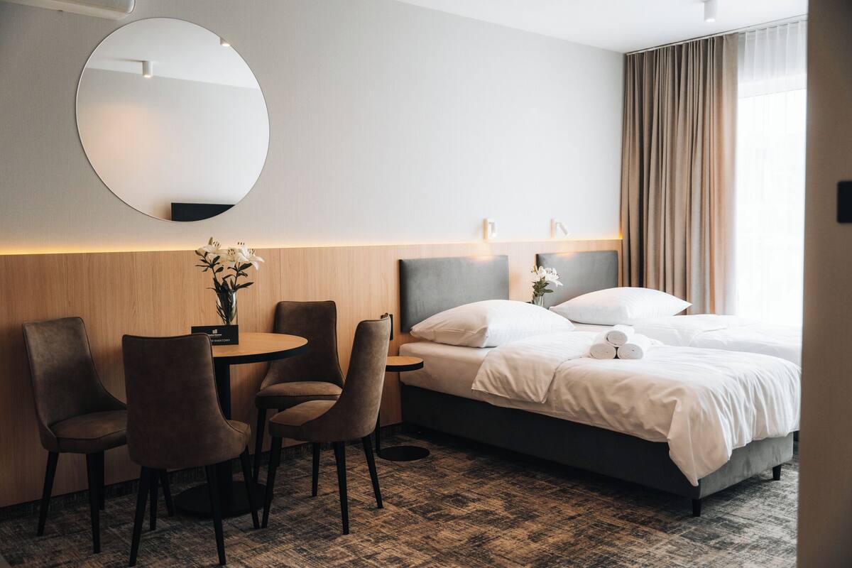 A hotel room with a bed, two chairs, a small table, a round mirror, and a vase with flowers.