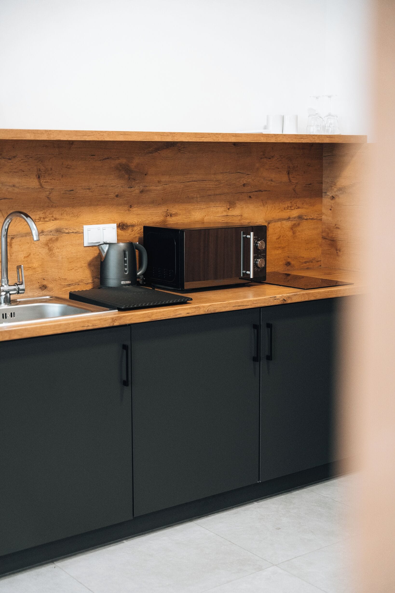 A modern kitchen with a wooden countertop, a black microwave, a kettle, and a sink.