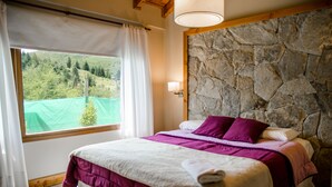 A bedroom with a stone wall, a bed with a purple and white bedspread, a window with a view of greenery, and a lamp on the wall. - MOUNTAIN CABIN DREAM (San Carlos de Bariloche)