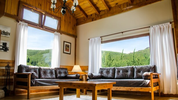 A wooden-floored living room with a leather sofa, a wooden coffee table, and large windows offering a view of green hills. - MOUNTAIN CABIN DREAM (San Carlos de Bariloche)