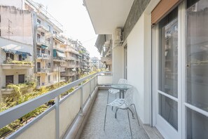 Uma varanda com uma mesa de vidro e duas cadeiras, com vista para uma fileira de edifícios altos com varandas.