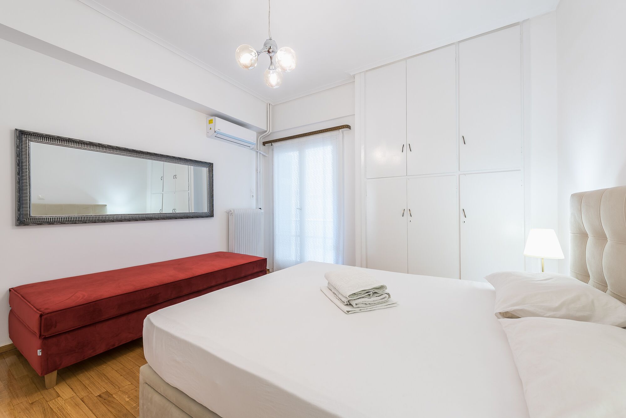 Une chambre à coucher avec un grand lit, un banc rouge, un miroir et des armoires blanches.