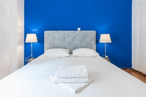 A neatly made bed with white linens and a folded towel, two white pillows, and a grey upholstered headboard.