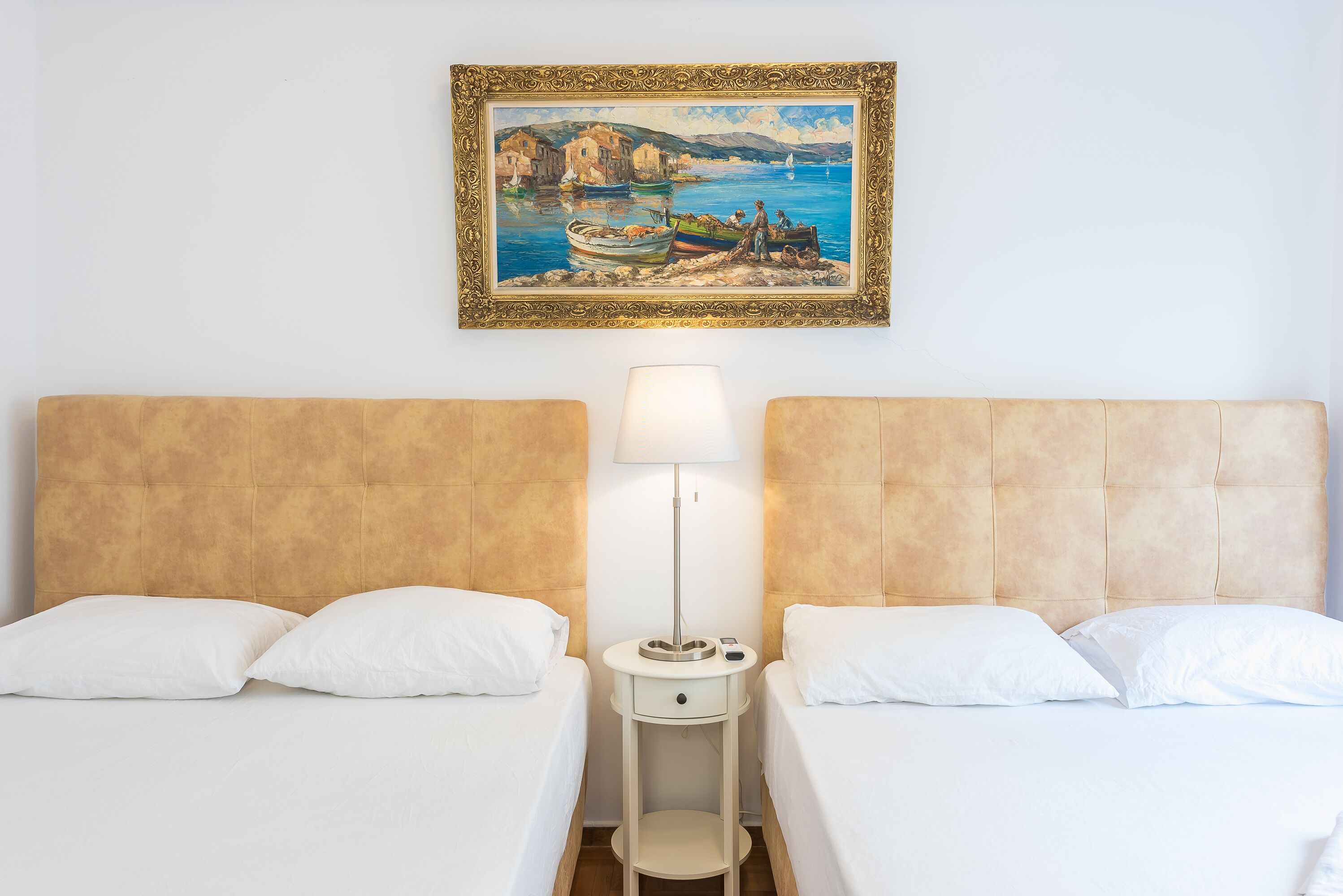 A hotel room with two beds, a nightstand with a lamp, and a framed painting of a coastal scene.