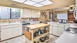 Une cuisine avec des meubles blancs, un îlot central en bois, un four encastré et une fenêtre avec des stores.