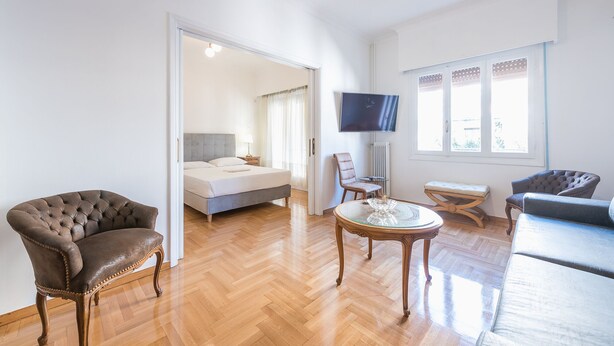 A spacious living room with a large bed, a TV, and a seating area with a table and chairs. - Victoria Supreme Suite (Athens)