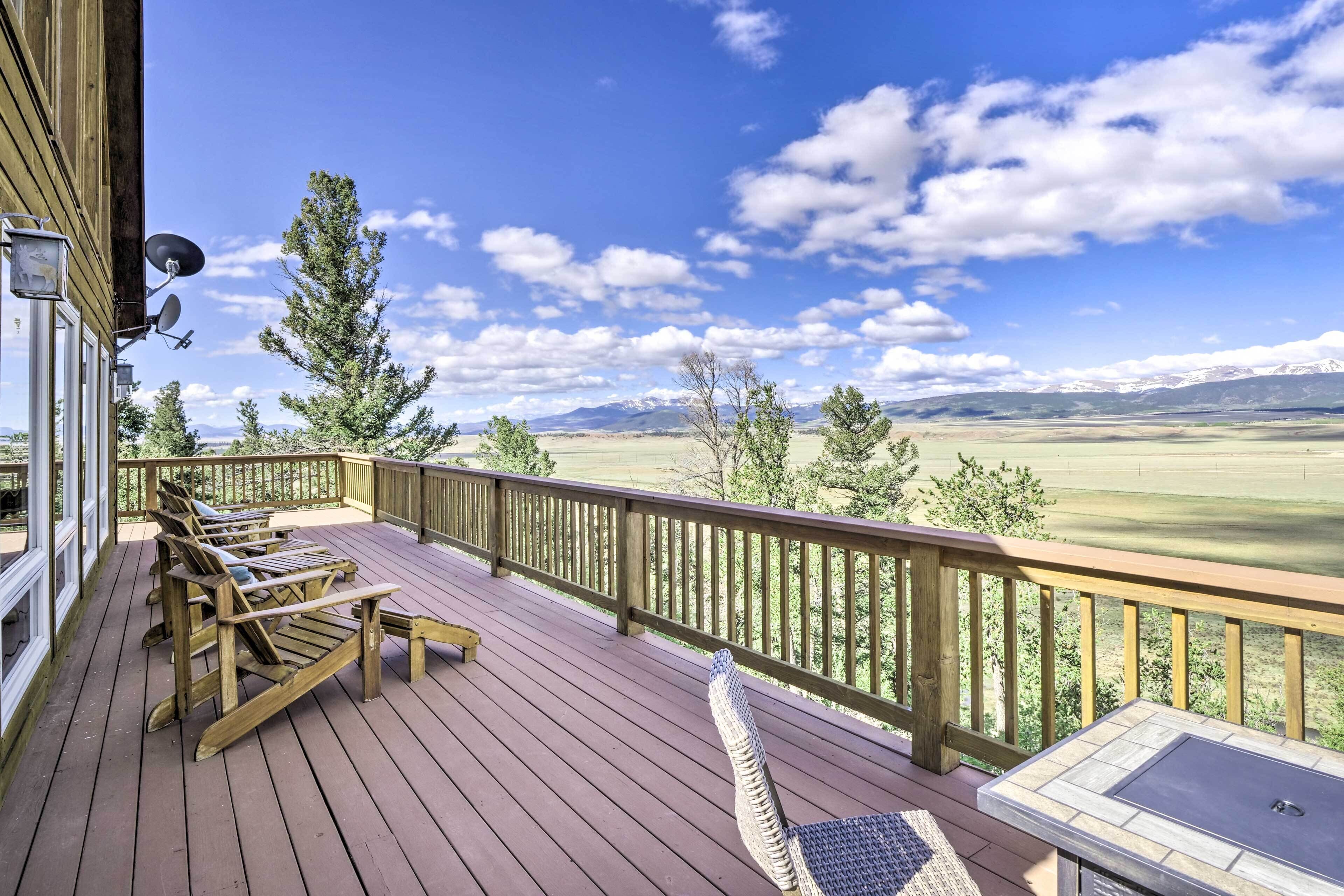 Une terrasse en bois, dotée de mobilier d’extérieur et d’une cheminée en pierre, qui offre une vue imprenable sur un paysage magnifique, avec des montagnes et des arbres.