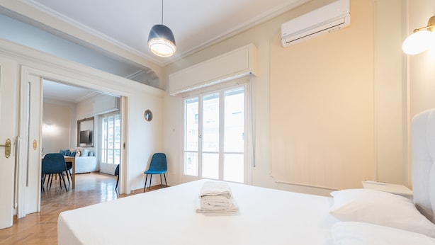 A hotel room with a large bed, a desk, and a chair. There is a window with blinds, an air conditioning unit, and a wall-mounted TV. - Athens Empire Paradise Apartment (Athens)