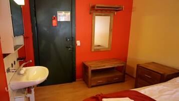 A small room with a white sink, a wooden bench, a mirror, and a bed with a red blanket.