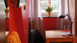 Una habitación con escritorio de madera, una silla negra, pared roja, ventana con planta en maceta y una lámpara.