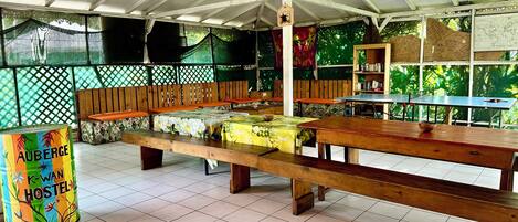 A hostel with wooden benches and tables, a tiled floor, and a green backdrop.