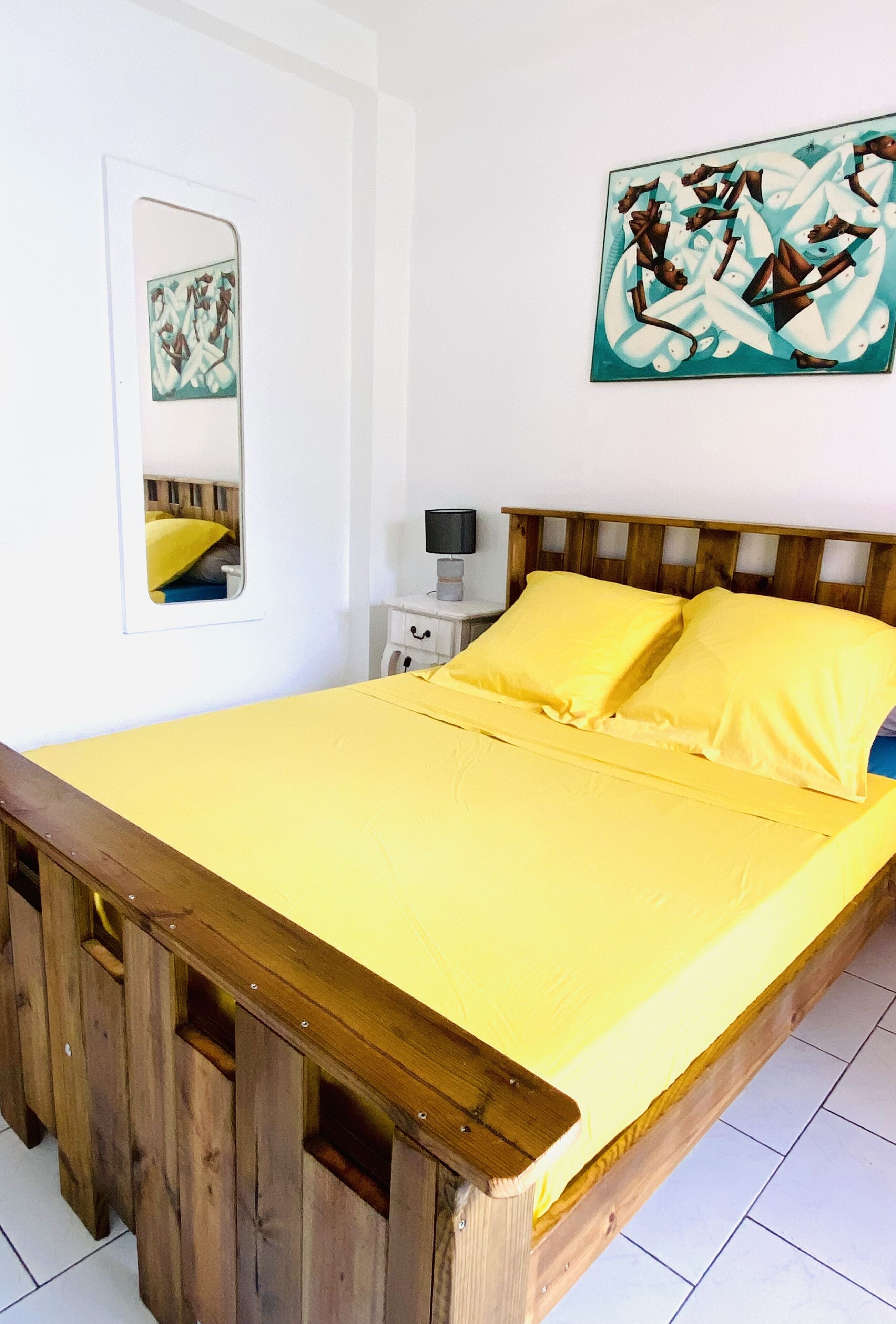 A room with a wooden bed frame, yellow bedding, a white wall, a small bedside table with a lamp, and a painting above the bed.