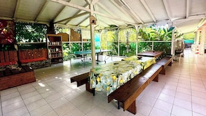 An indoor dining area with a long table covered in a floral tablecloth, benches, and a bookshelf. - Auberge K-Wan Hostel (Deshaies)