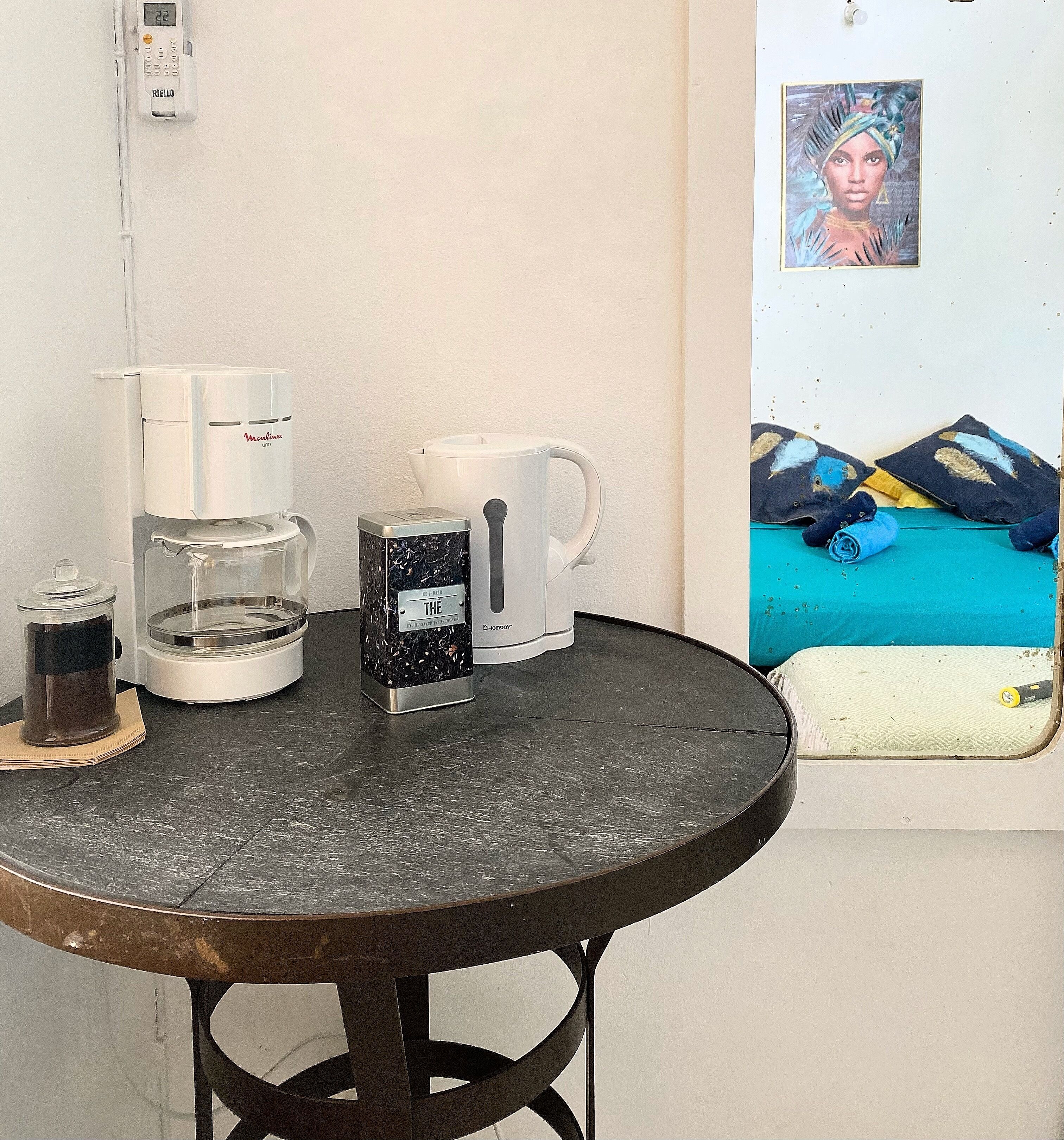A small round table with a coffee maker, a mug, and a thermos. A picture of a bed with pillows and a blanket is on the wall behind the table.