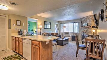 A kitchen with a central island, wooden cabinets, and a dishwasher. There is a dining area with a table and chairs, and a living room with a sofa and a coffee table.