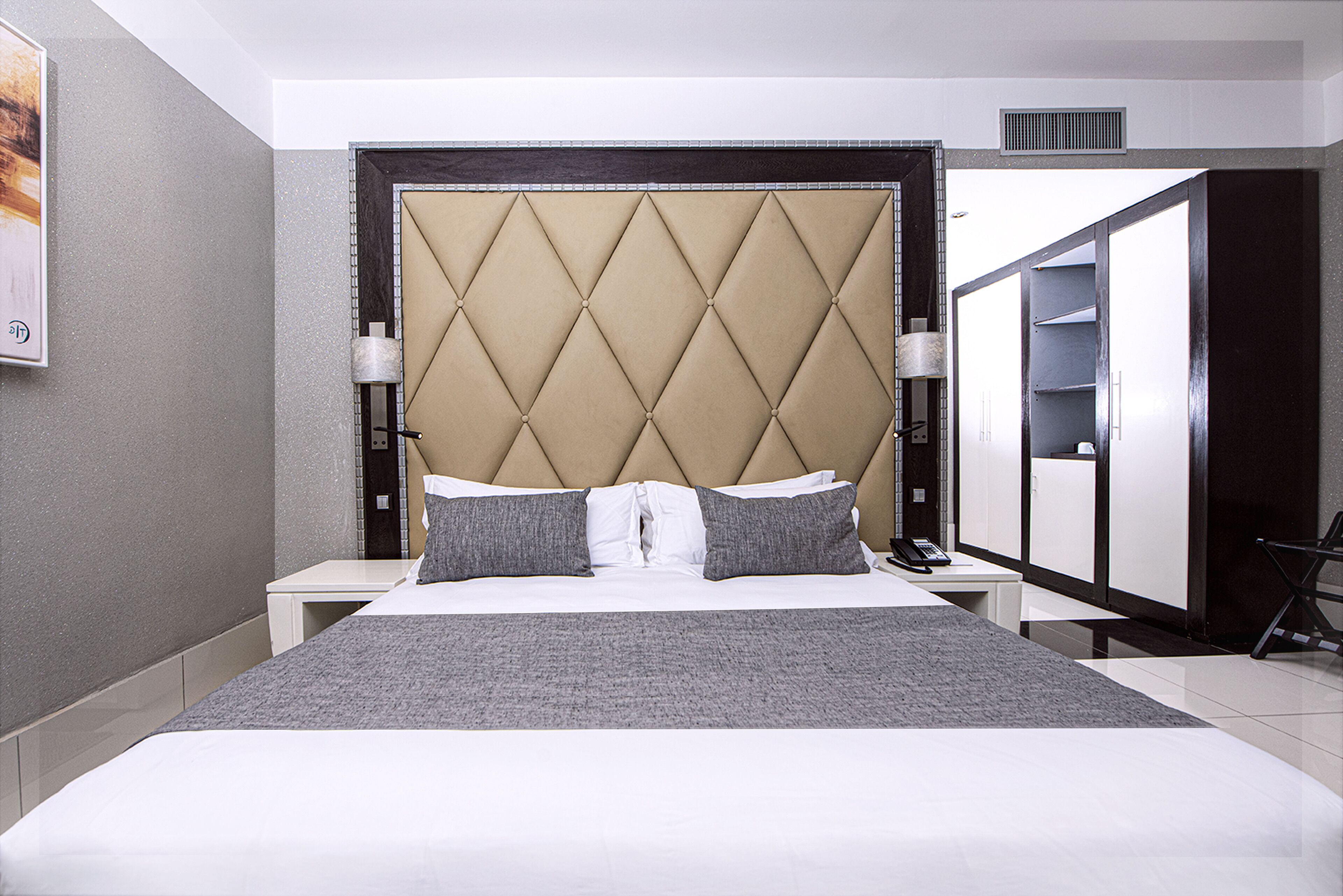 A hotel room with a large bed, a headboard with a diamond pattern, two bedside tables with lamps, and a wardrobe.