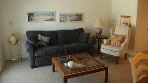 Een woonkamer met een donkergrijze bank, een glazen salontafel, een beige fauteuil en twee ingelijste foto's aan de muur.