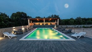 A rectangular swimming pool with a wooden deck, surrounded by lounge chairs and a pergola with lights. - Apartments Karen (Marina)