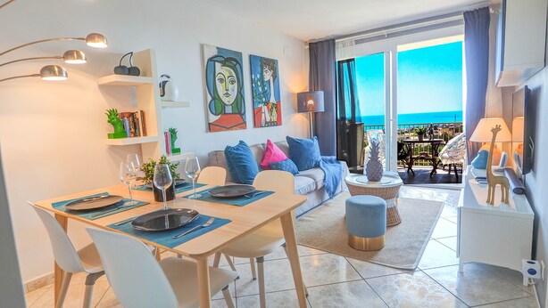 Un salon moderne avec un coin repas, un canapé agrémenté de coussins colorés, un balcon avec vue et des œuvres d’art accrochées au mur.