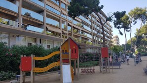 Ein Spielplatz mit Rutsche und Klettergerüsten, umgeben von einem Gebäude mit mehreren Balkonen und einem Garten mit rosa Blumen.