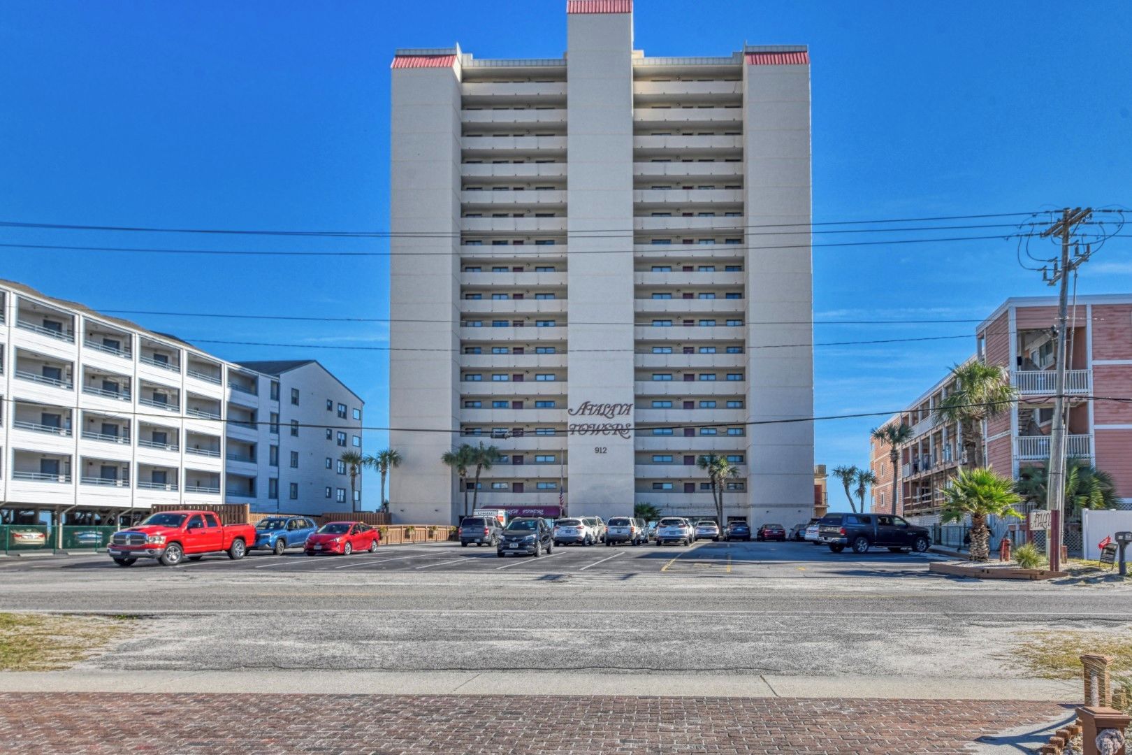 Un hôtel à plusieurs étages, avec une enseigne bien visible sur la façade, un parking à l’avant et un ciel bleu dégagé.