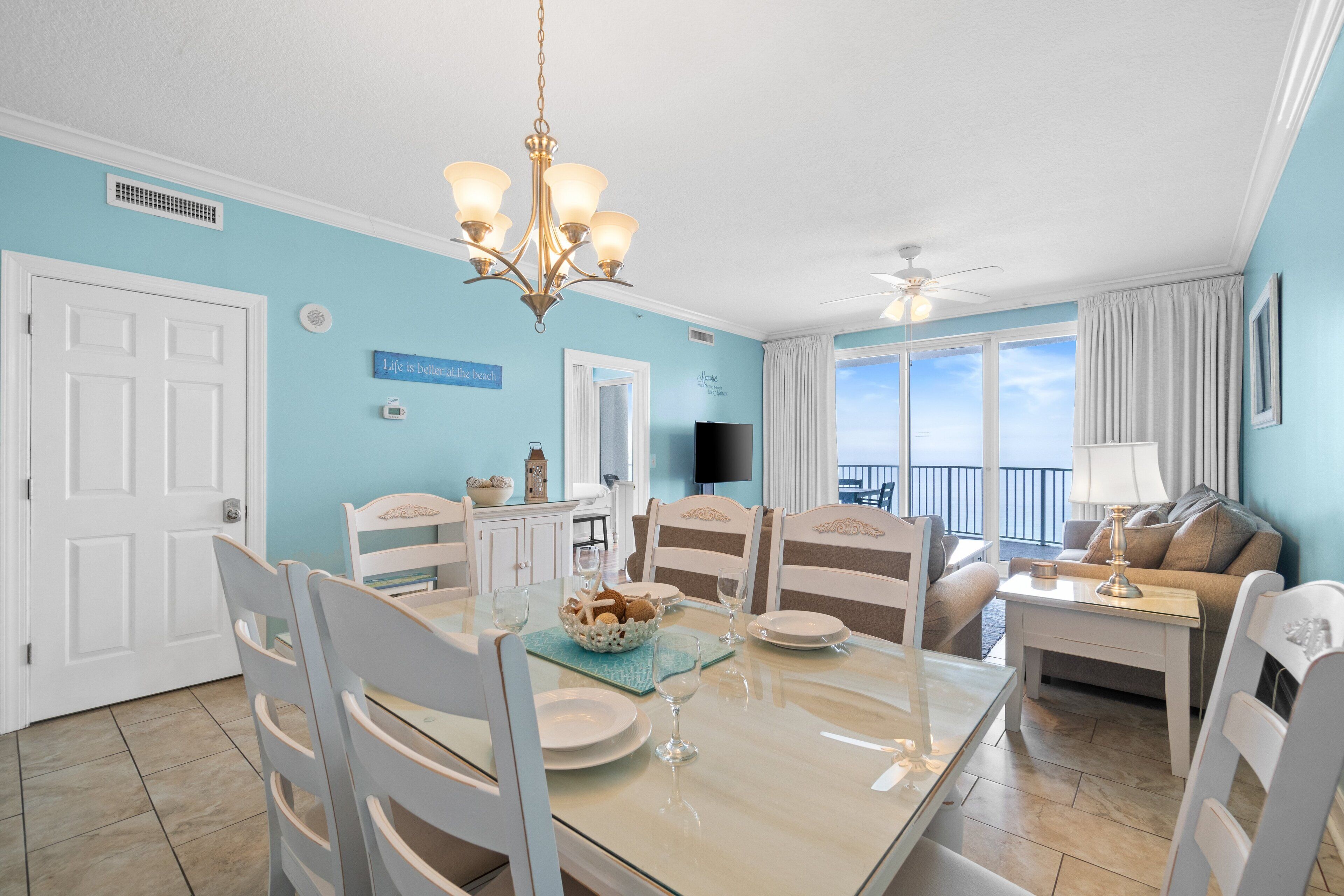 A dining area with a table set for four, surrounded by white chairs. A chandelier hangs above the table, and a television is mounted on the wall.