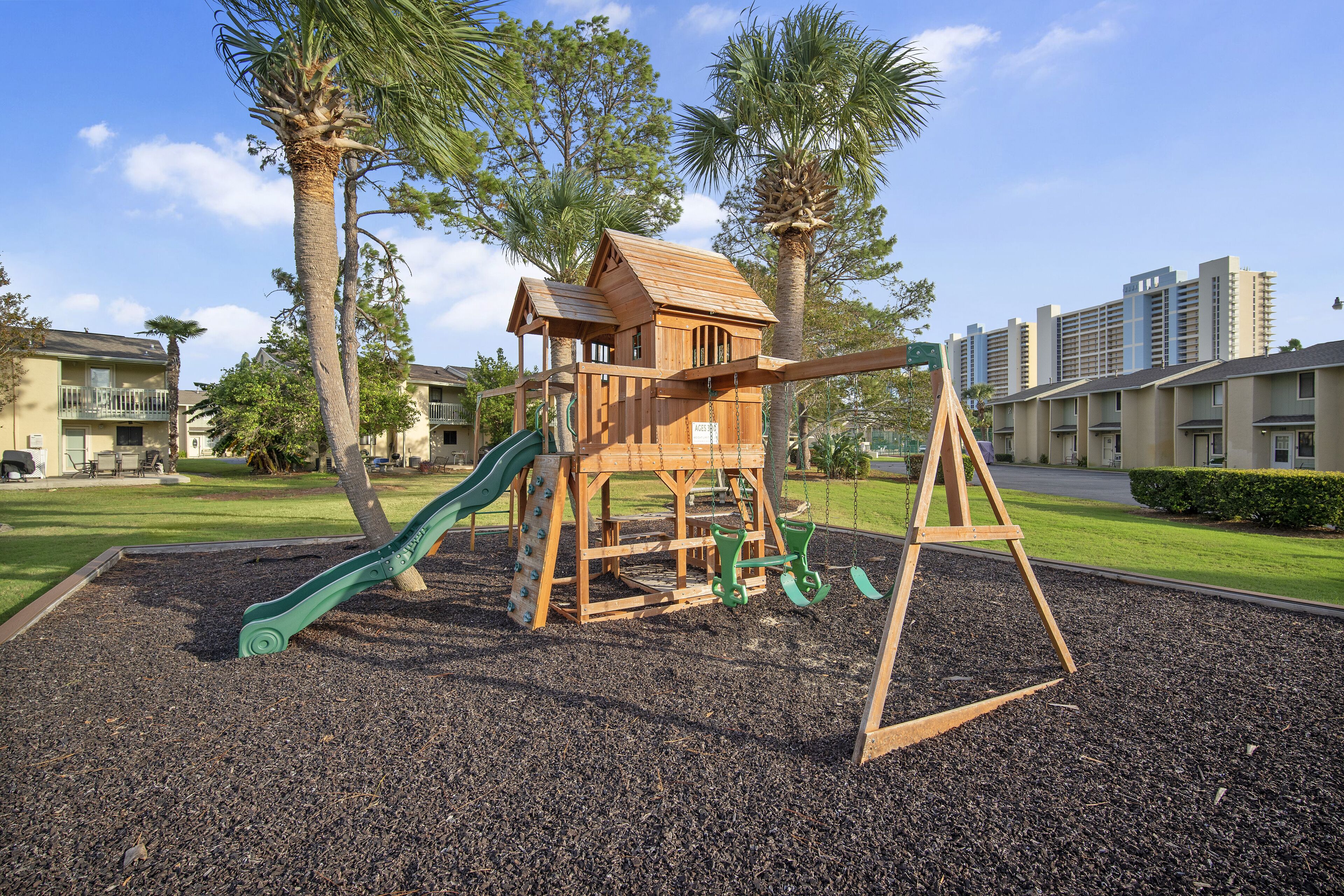 Estructura de juego de madera con tobogán y columpios, rodeada de palmeras y una zona de juegos.