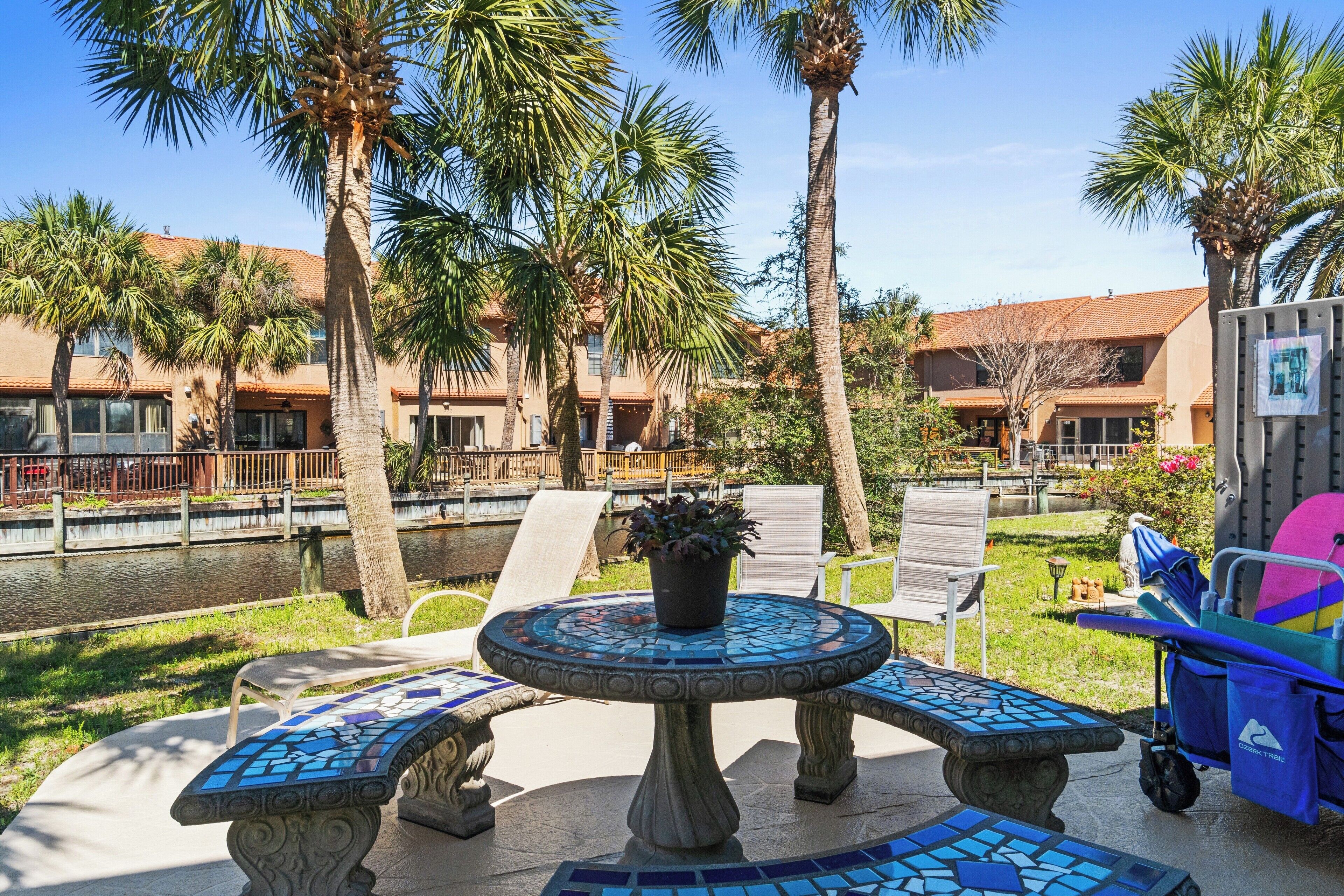 A patio with a mosaic table and benches, palm trees, and a pool.