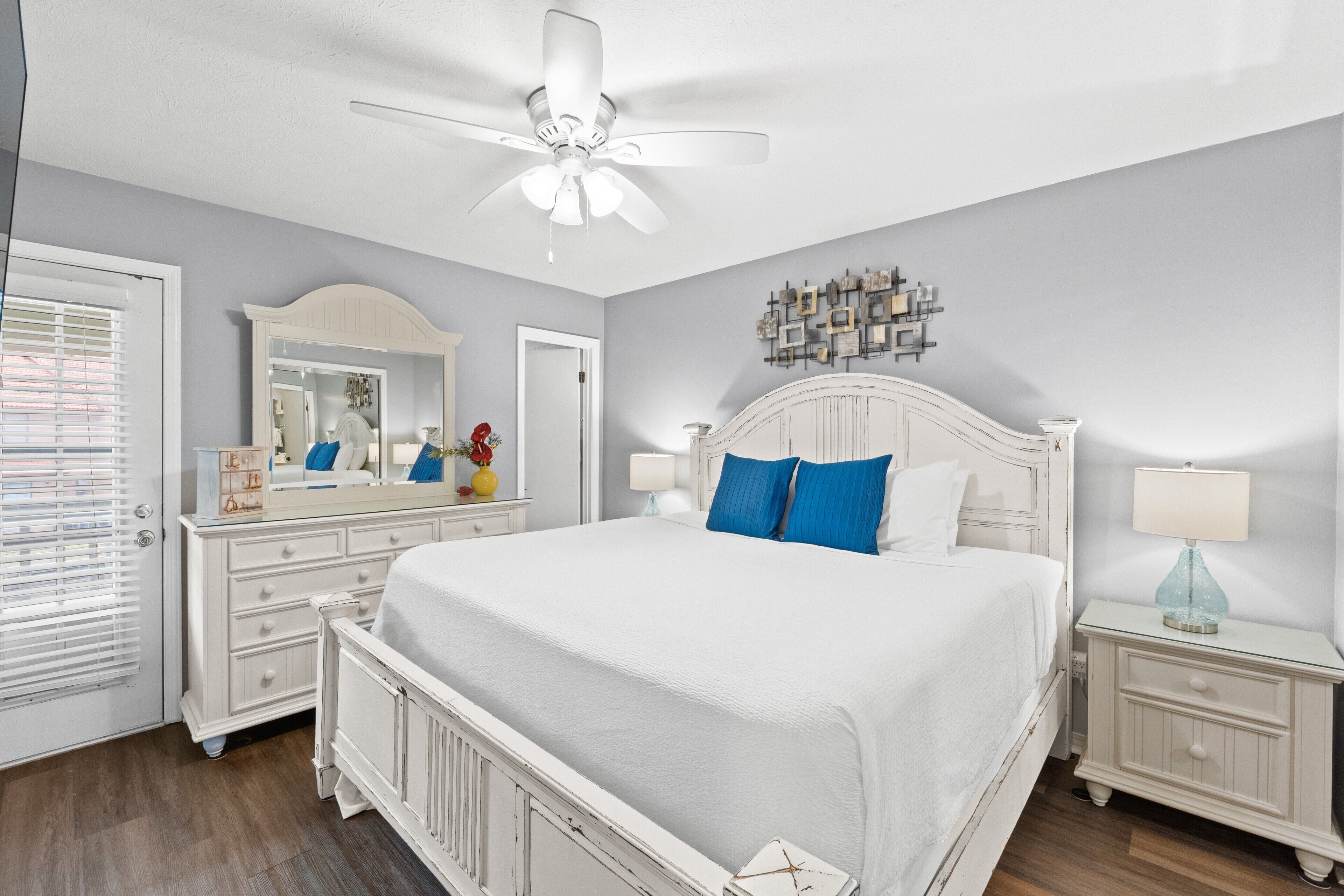A bedroom with a white bed, a ceiling fan, a dresser, and a nightstand with a lamp.