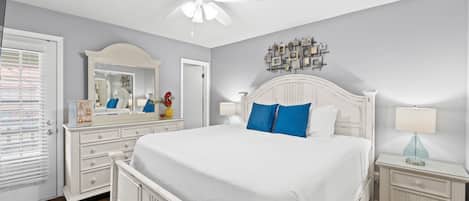 A bedroom with a white bed, a ceiling fan, a dresser, and a nightstand with a lamp.