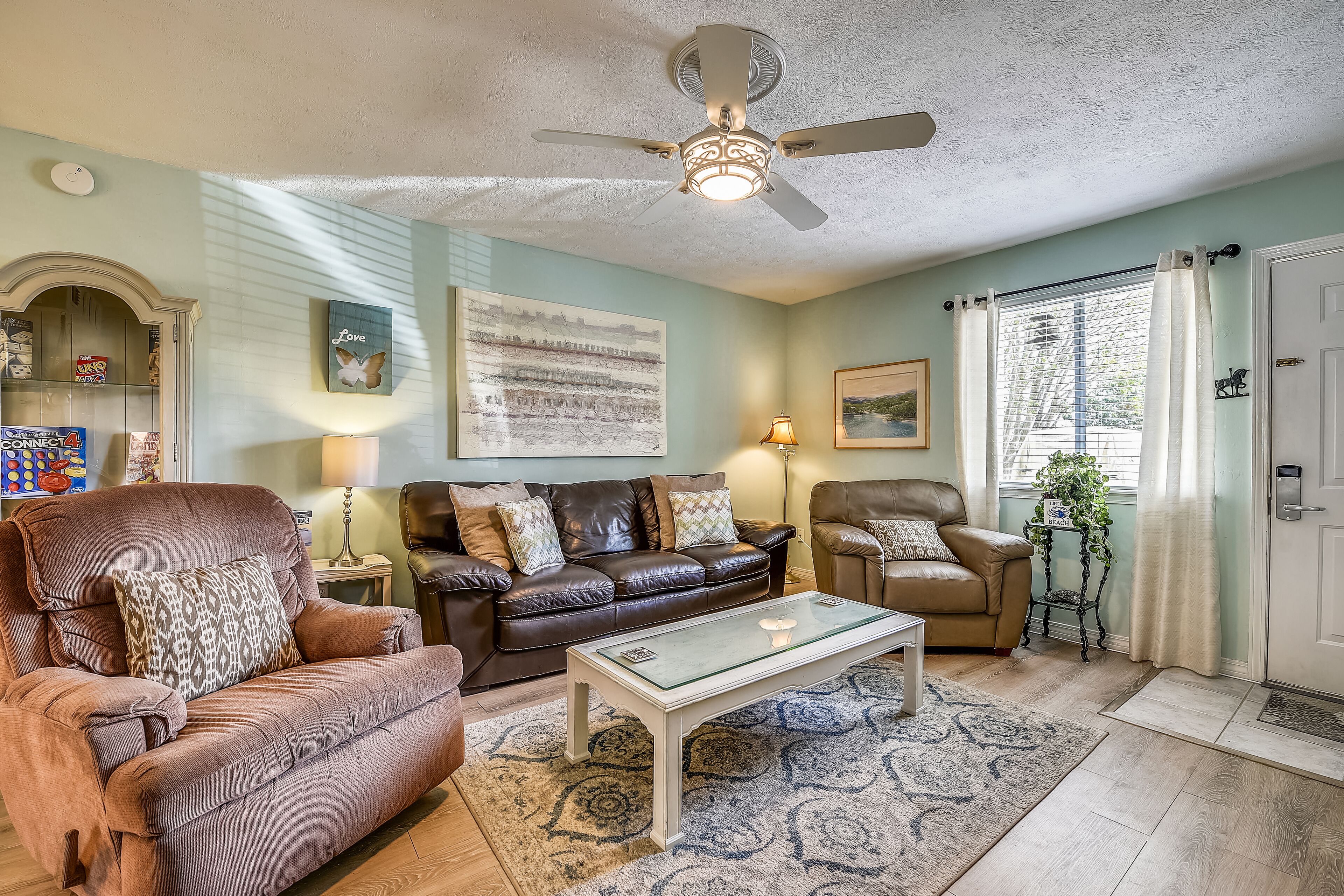 A living room with a ceiling fan, a brown leather sofa, a glass coffee table, and a patterned rug.
