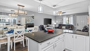 Eine moderne Küche mit weißen Schränken, einer Granit Arbeitsplatte und Pendelleuchten. Es gibt einen Essbereich mit weißen Stühlen und einem Tisch sowie eine Wohnbereich mit einer Couch und Sesseln.