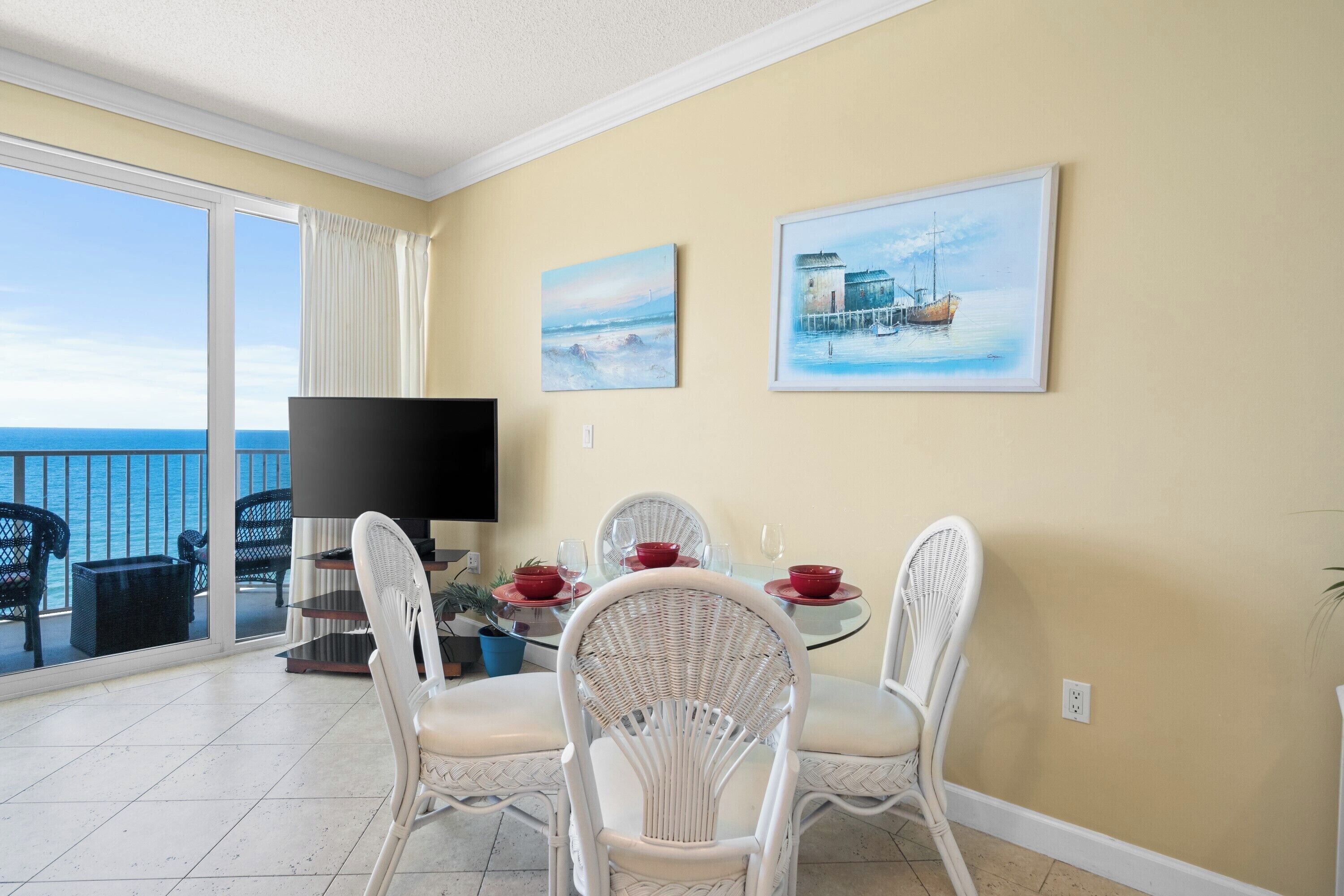 A dining area with a balcony overlooking the sea, featuring a table set for two with white wicker chairs, a flat-screen TV, and artwork on the wall.
