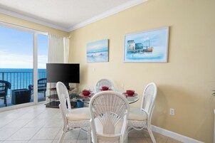 A dining area with a balcony overlooking the sea, featuring a table set for two with white wicker chairs, a flat-screen TV, and artwork on the wall.