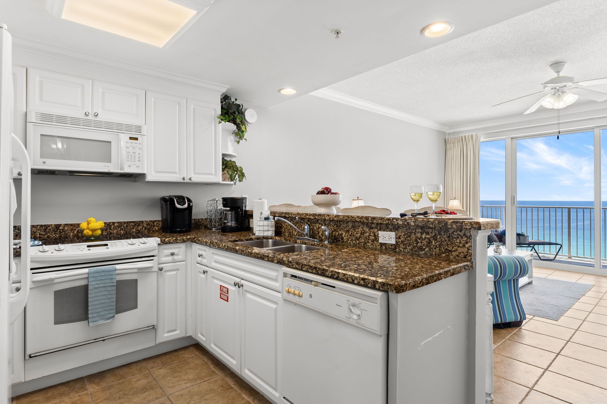 A modern kitchen with white cabinets, granite countertops, a built-in oven, and a dishwasher. There is a large window offering a view of the ocean.