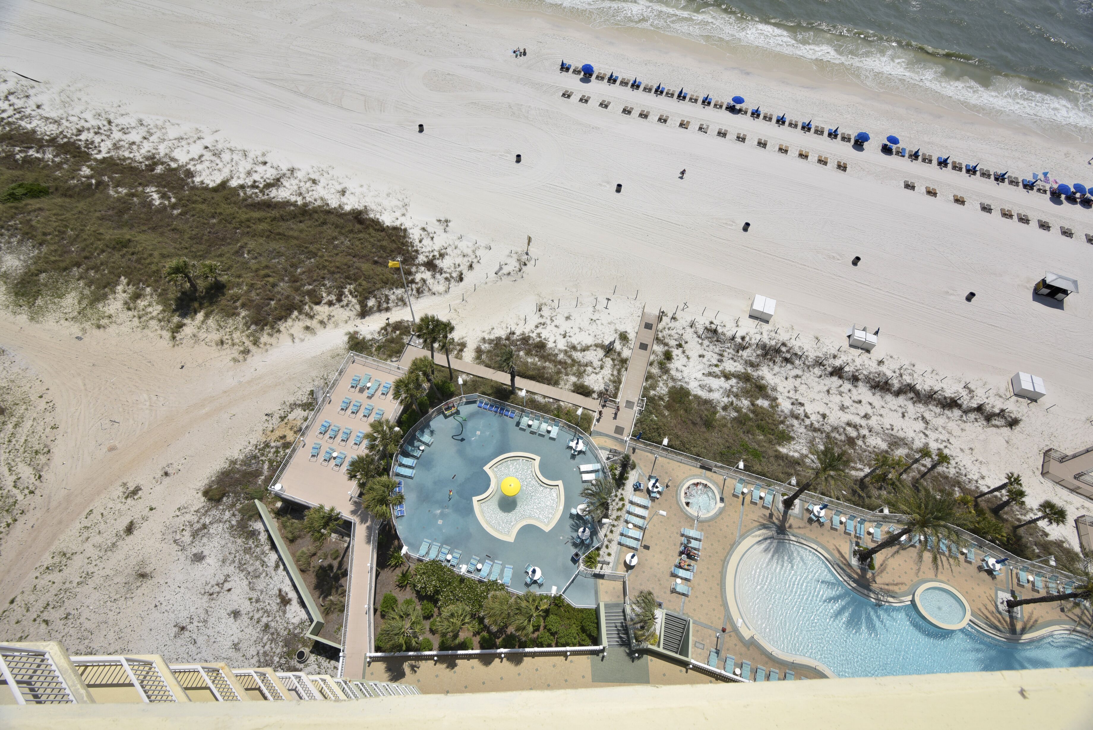 Vue aérienne d’un complexe hôtelier de bord de mer, avec une piscine, des chaises longues et une plage aménagée avec des parasols.