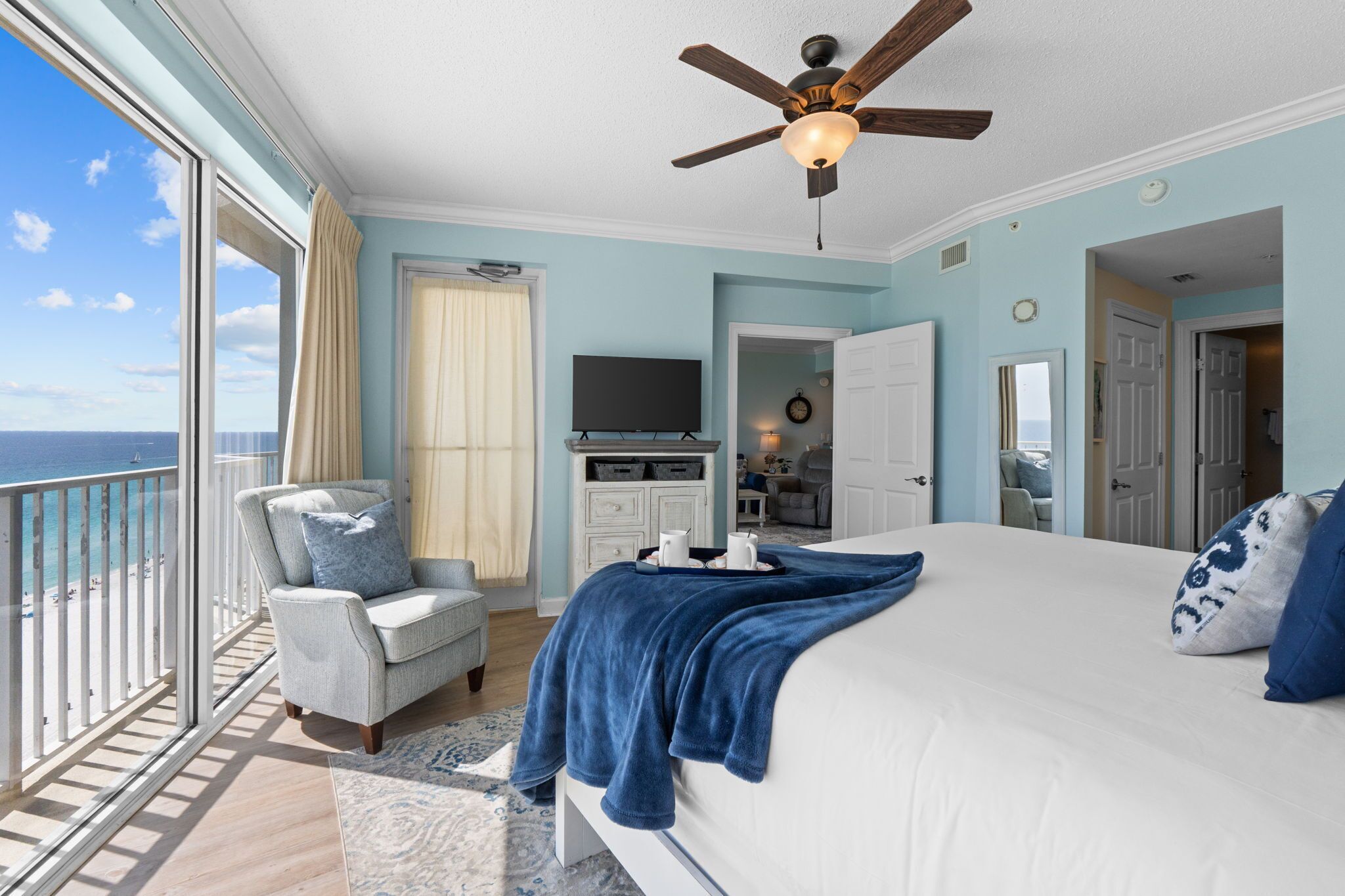 A bedroom with a bed, a chair, a TV, a ceiling fan, and a view of the ocean.