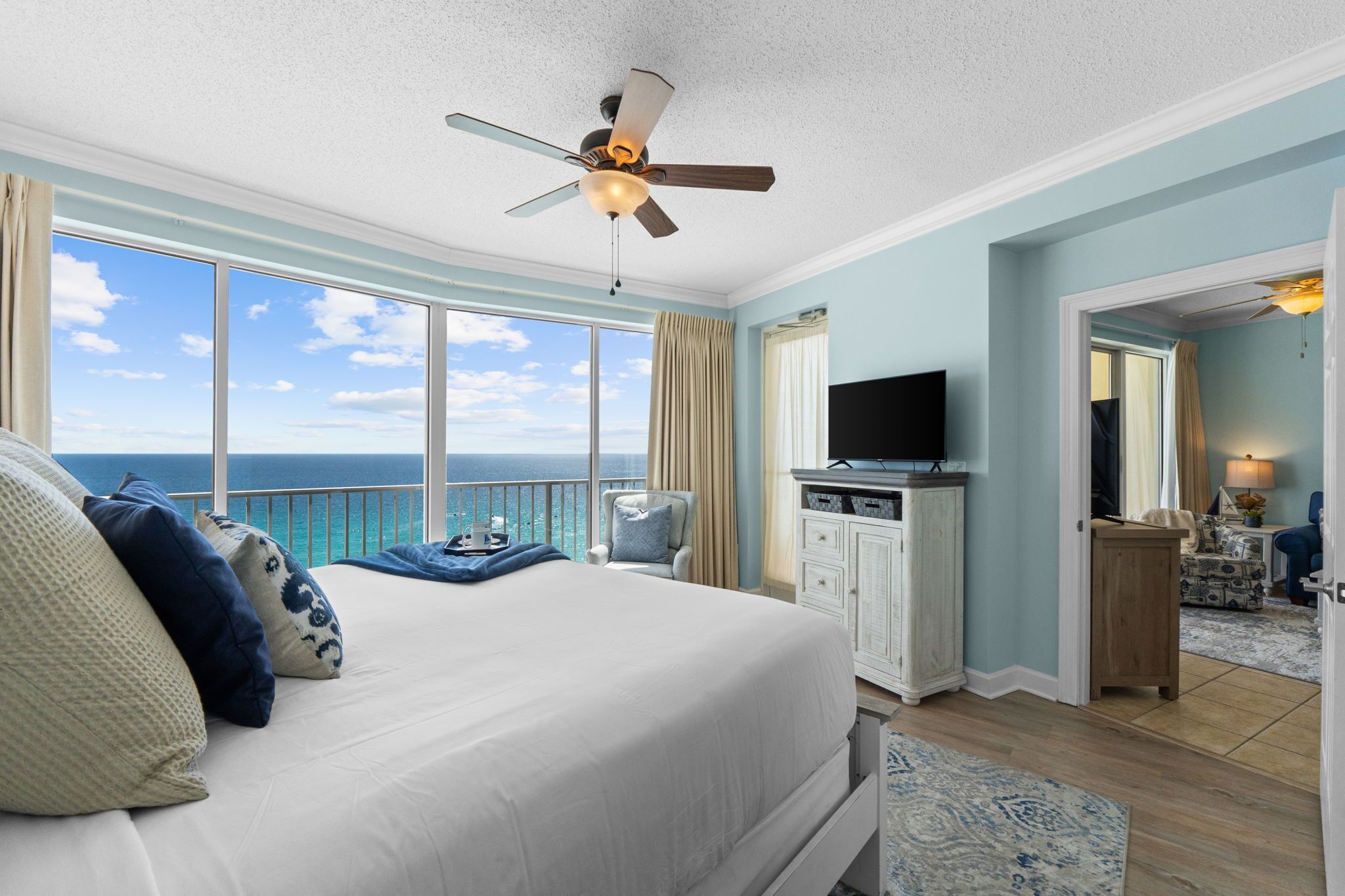 A bedroom with a bed, a TV, a ceiling fan, a view of the ocean, and a door leading to another room.