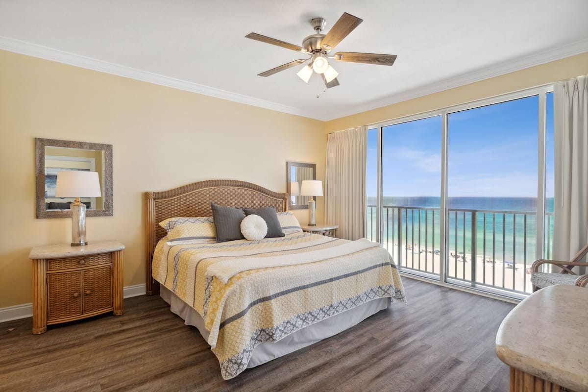 Un dormitorio con una cama grande, ventilador de techo, mesita de noche, lámpara, ventana con vista a la playa y balcón con una silla.