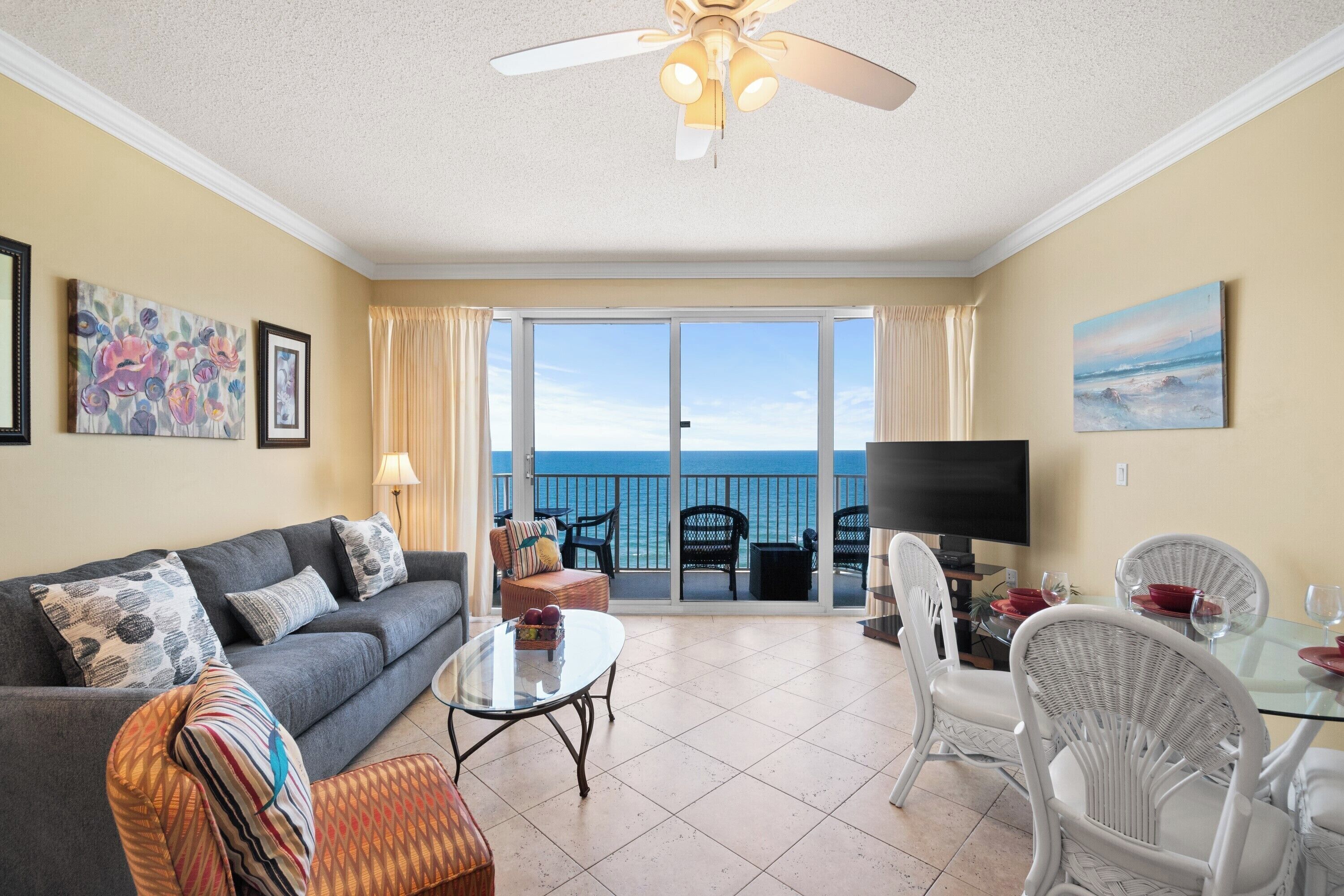 A living room with a sofa, armchairs, a coffee table, and a dining table set for two. A ceiling fan is visible, and there are large windows offering a view of the ocean.