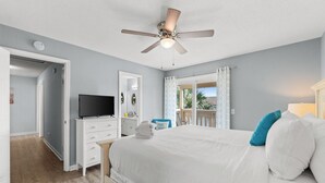 A bedroom with a bed, a ceiling fan, a television, a desk, and a window with curtains.