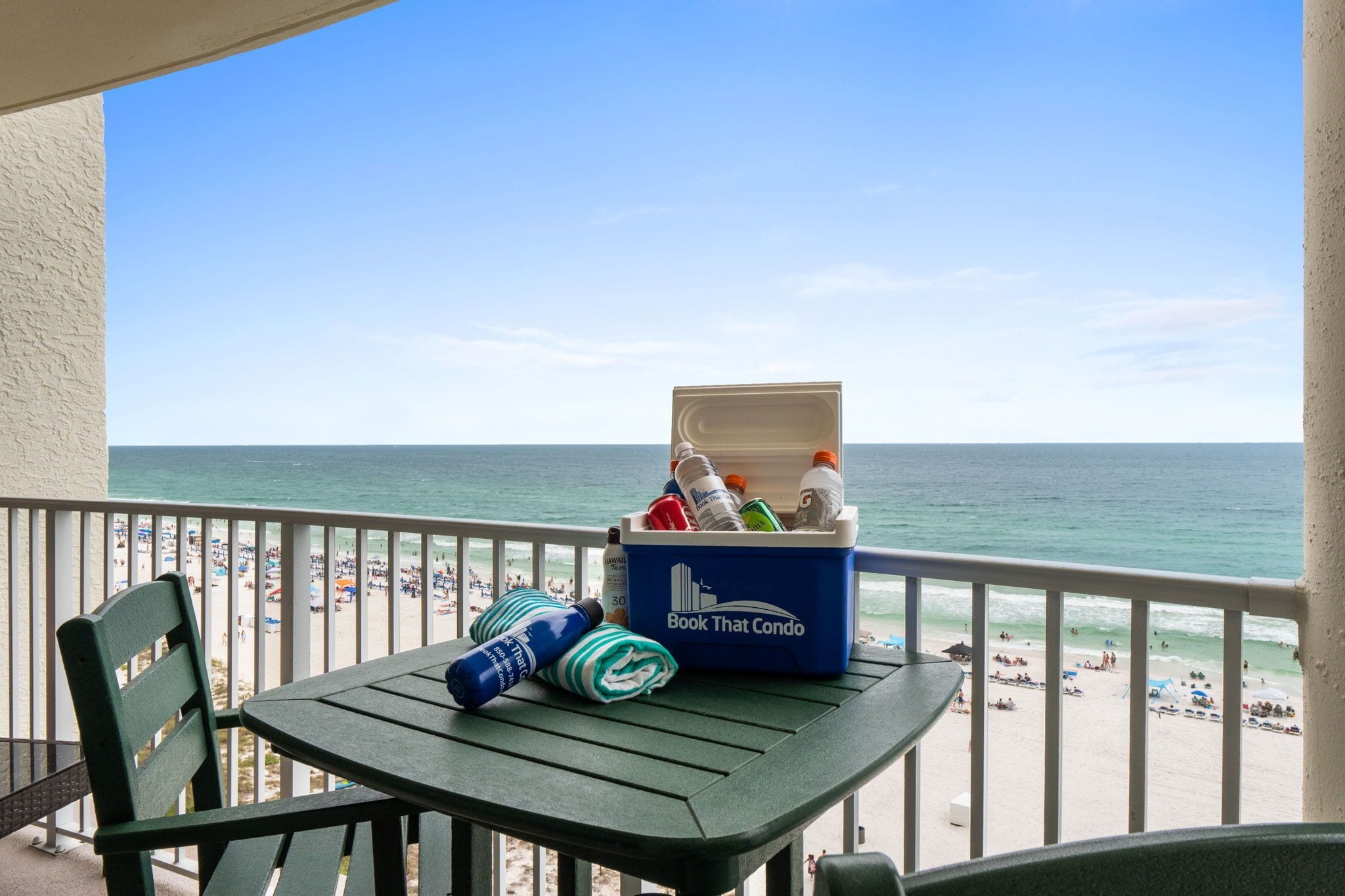 A balcony with a table, a cooler, and a chair overlooking a beach with people and umbrellas.