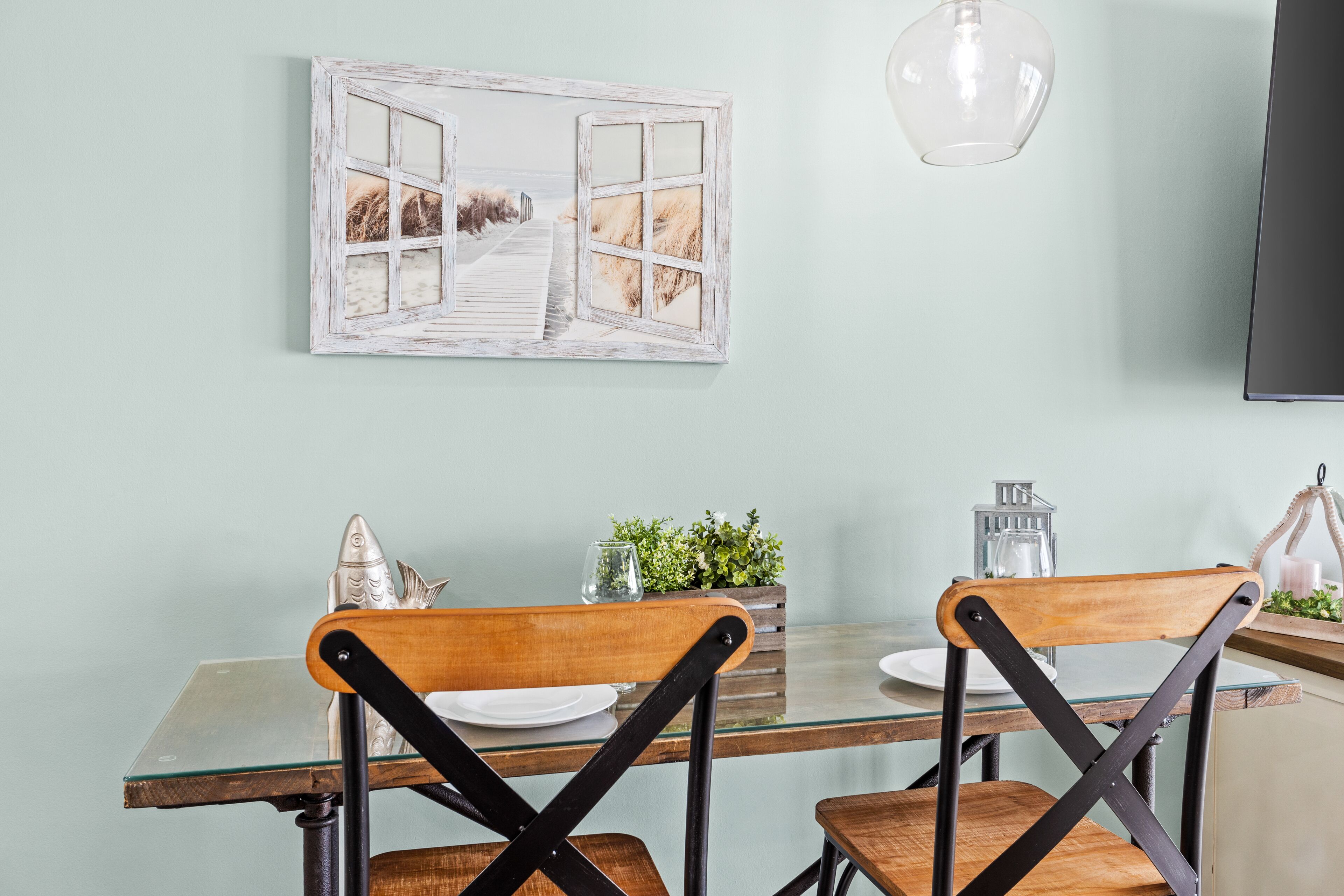 Un coin repas avec une table en verre, des chaises en bois, un tableau représentant une scène de plage et un luminaire suspendu.